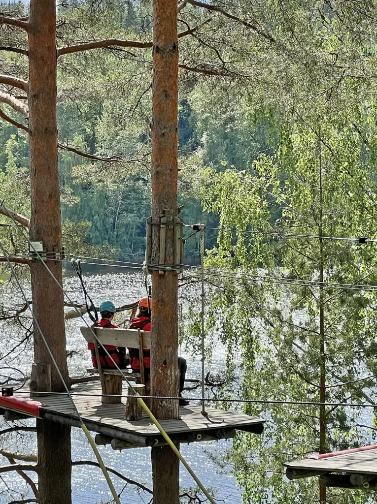 Kaksi seikkailijaa istuvat korkealla olevan tason päällä olevalla penkillä ja katsovat järvimaisemaa Leppävirralla sijaitsevassa seikkailupuistossa